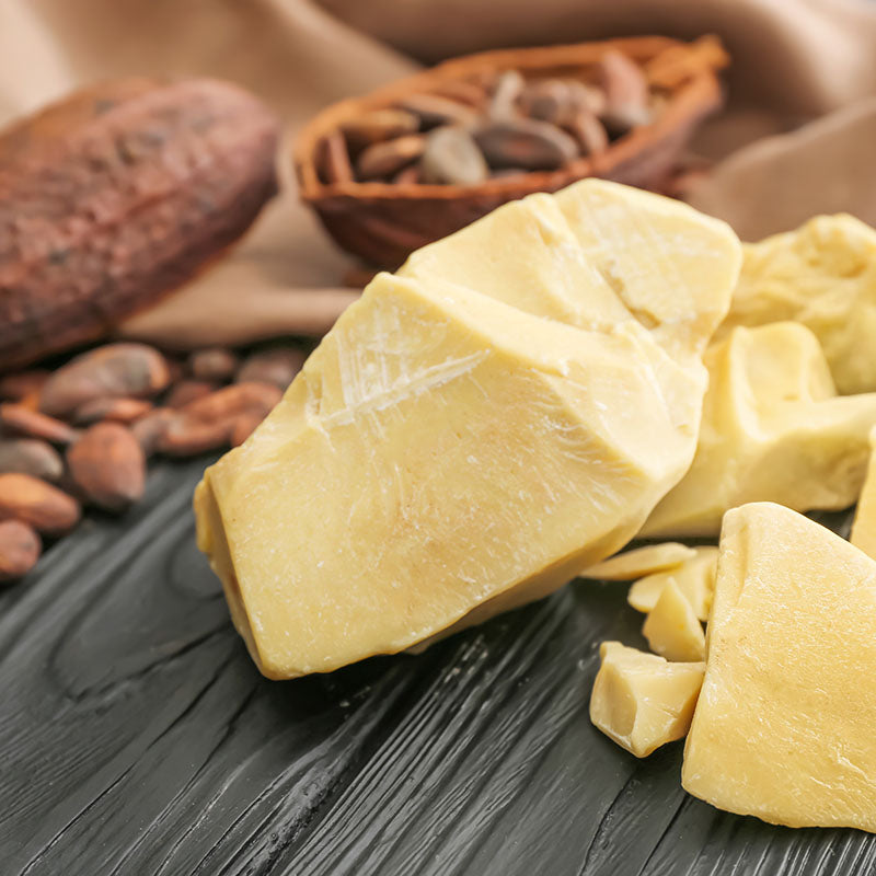 Chunks of yellow cocoa butter on a dark surface with cocoa beans and a wooden bowl in the background.