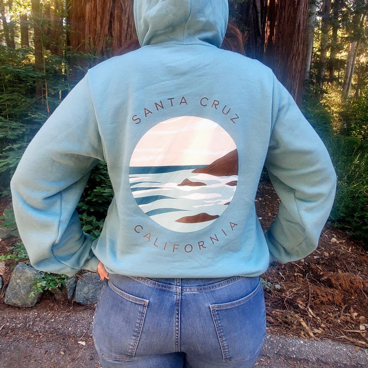 Person wearing a light blue hoodie with 'Santa Cruz California' design in a forest setting