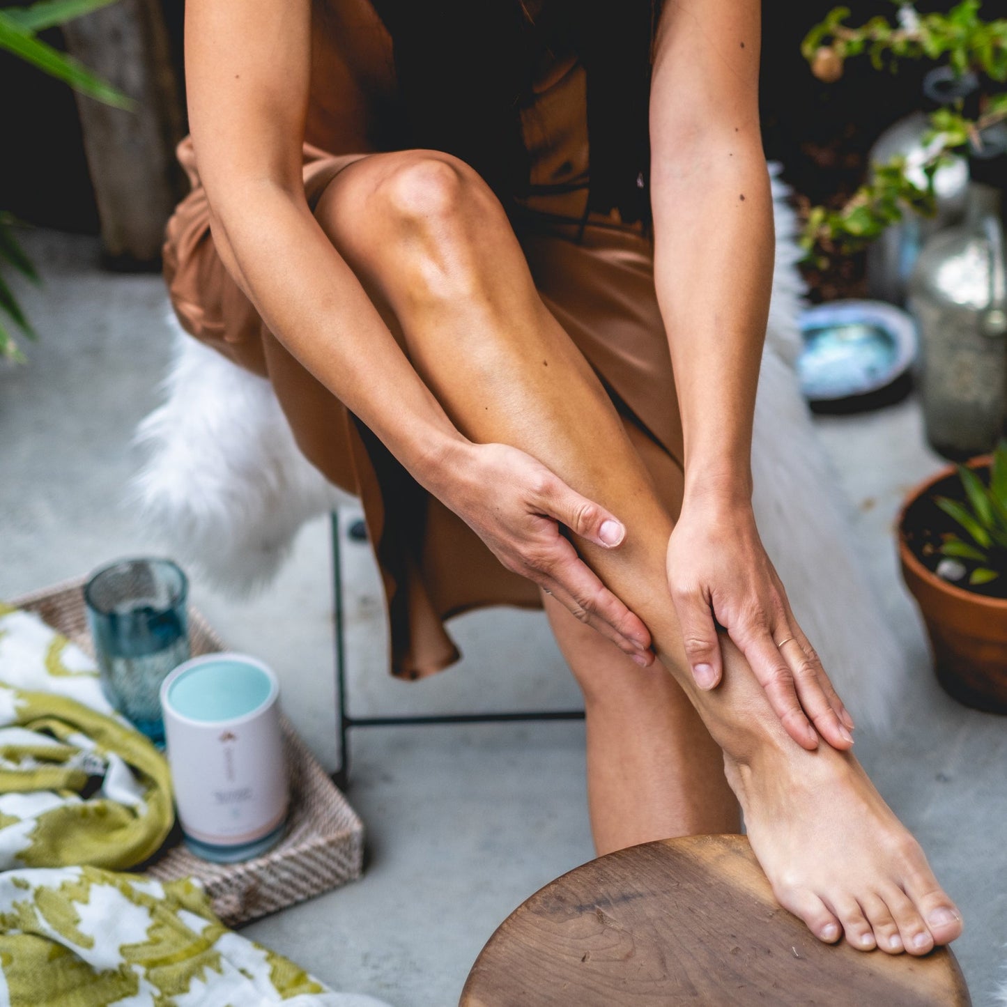 Person sitting on a stool, massaging their foot in a home setting with plants and a candle.