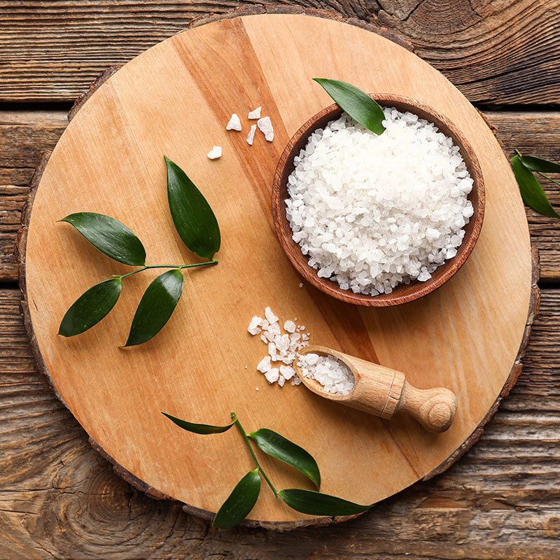 Wooden cutting board with a bowl of white salt, a wooden scoop, and green leaves on a rustic wooden surface.