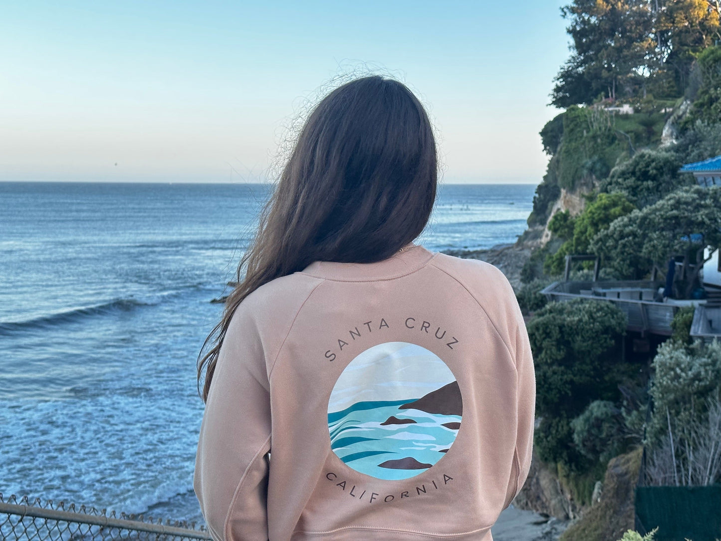 Person wearing a pink sweatshirt with a graphic design of Santa Cruz, California, overlooking the ocean.