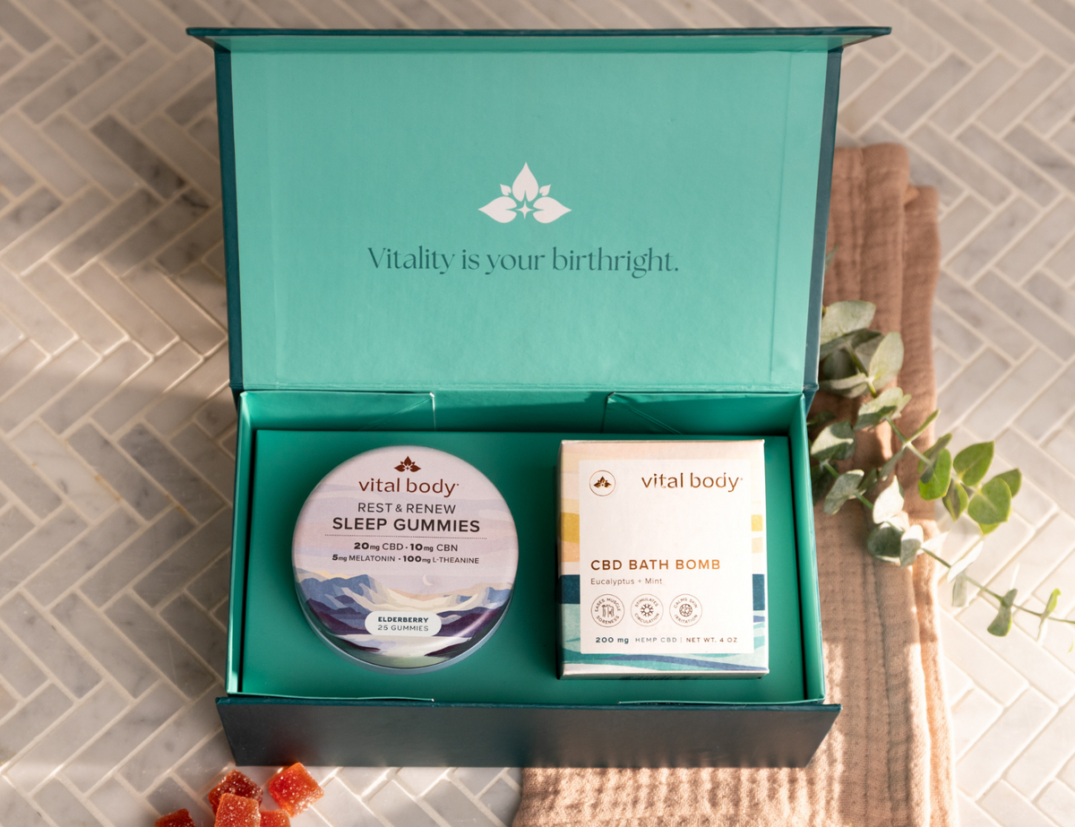 An open teal Vitality gift box on a light tiled surface, containing a round tin of sleep gummies and a boxed CBD bath bomb. A folded beige towel, eucalyptus sprigs, and a few orange gummy candies are arranged around the box.