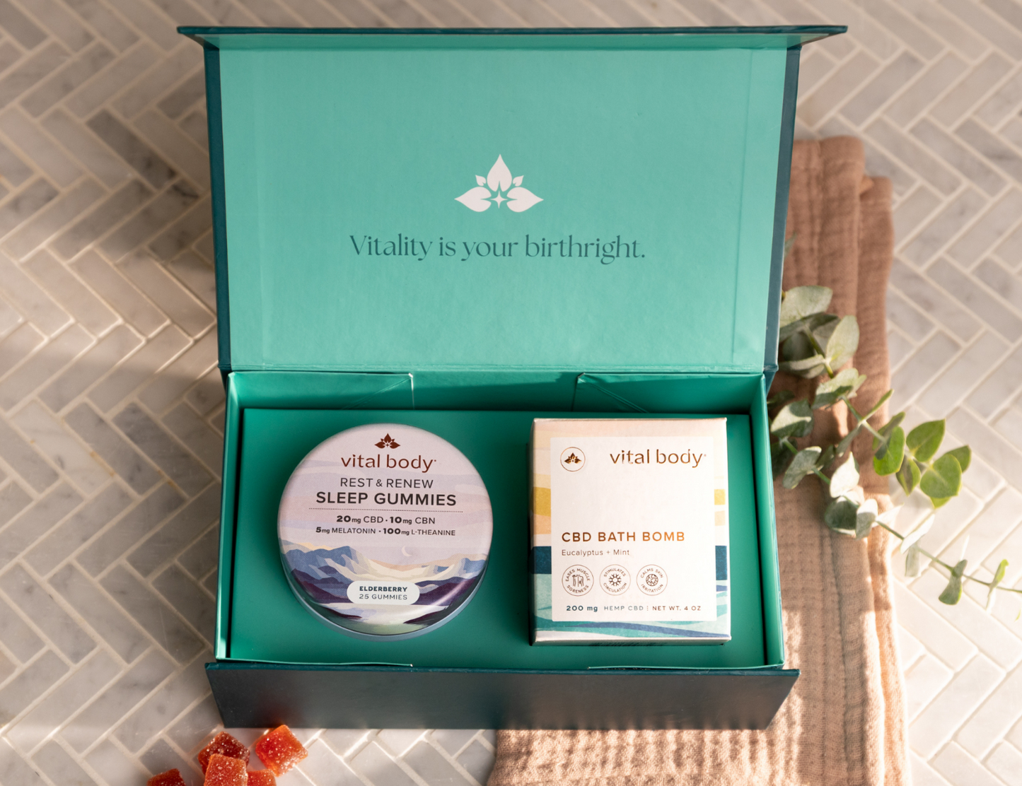 An open teal Vitality gift box on a light tiled surface, containing a round tin of sleep gummies and a boxed CBD bath bomb. A folded beige towel, eucalyptus sprigs, and a few orange gummy candies are arranged around the box.
