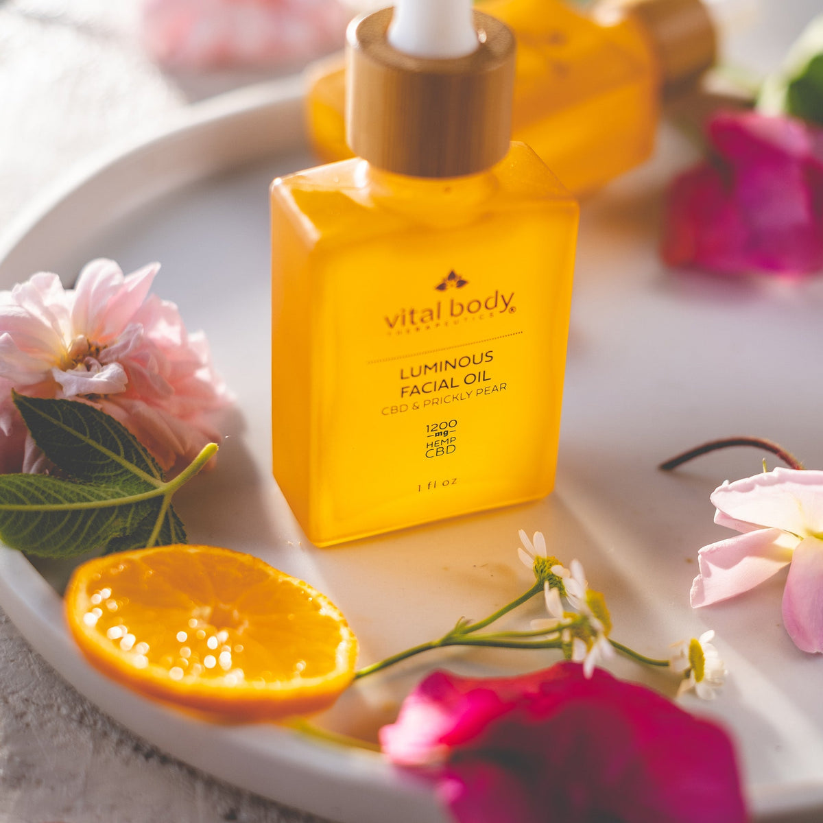 Vital Body Luminous Facial Oil bottle on a white plate with flowers and an orange slice.