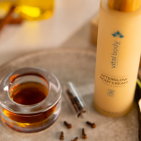 Vital Body Afterglow Body Cream bottle on a textured surface with a glass container and small bottles.