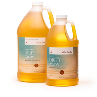 Two plastic gallon jugs of Vital Body Drift Away massage oil with a tropical blend scent, filled with golden yellow oil, on a white background.