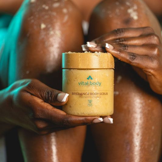 Person applying body scrub from a jar labeled 'vital body' to their skin.