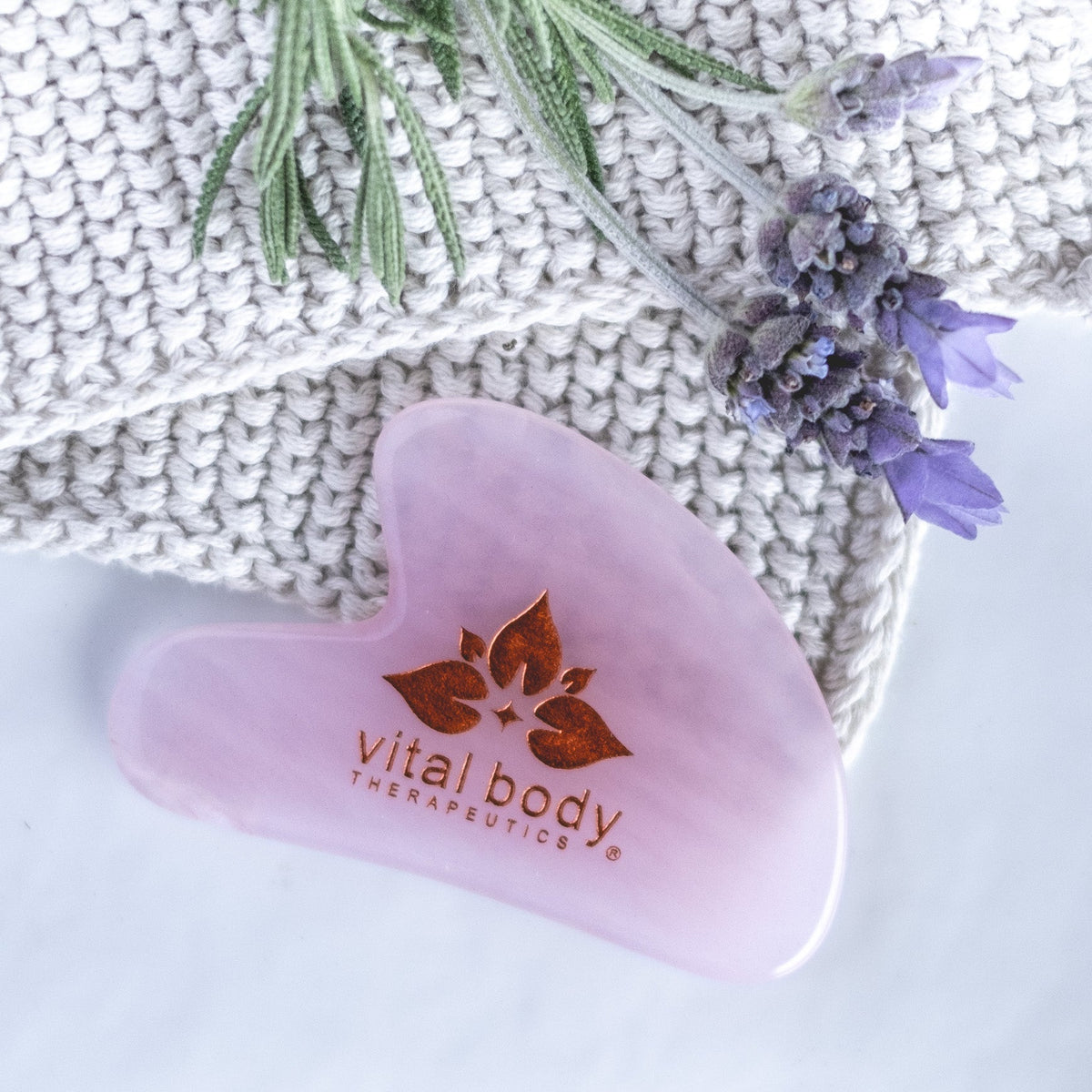 Pink heart-shaped tool with 'Vital Body Therapeutics' logo on a textured surface with lavender flowers.