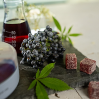 Frosted berries, a bottle of dark liquid, and cannabis leaves on a stone surface.