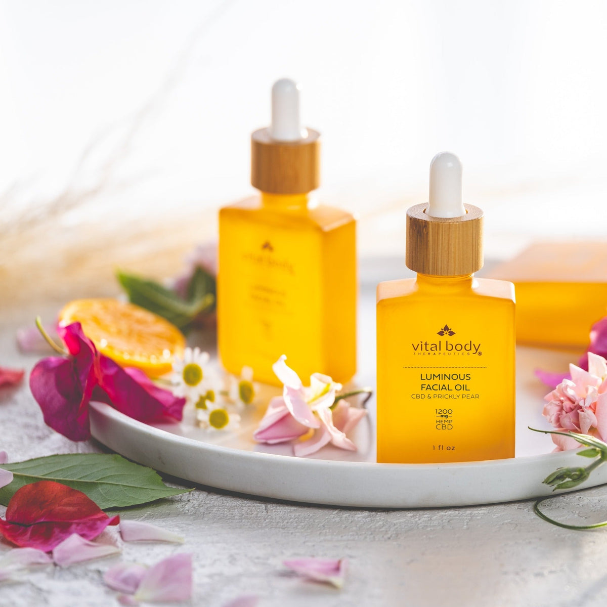 Two bottles of 'vital body' luminous facial oil on a white plate with flowers and fruits.