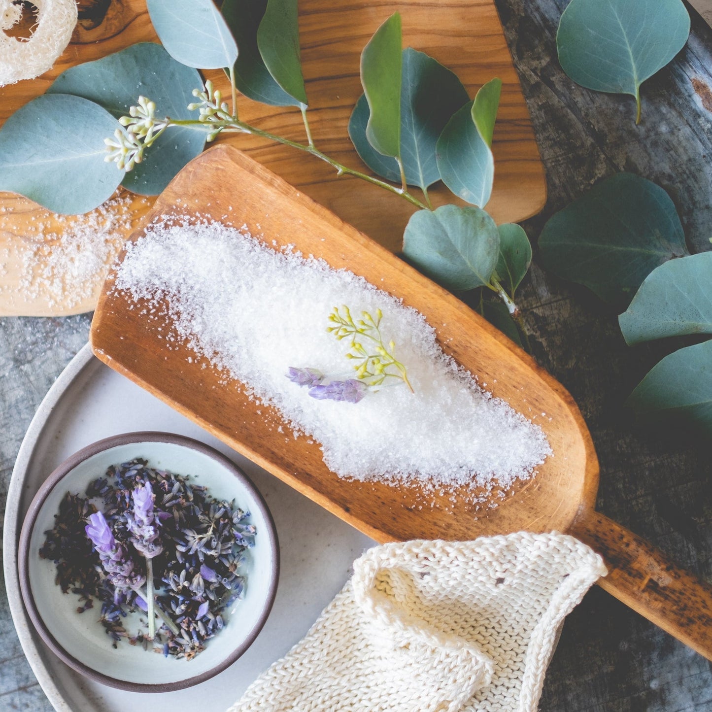 Wooden scoop with white powder, small bowl of dried lavender, and green leaves on a wooden surface.