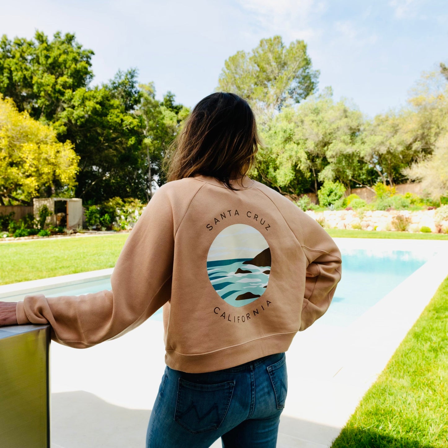 Person wearing a brown sweatshirt with a graphic design, standing by a pool with greenery in the background